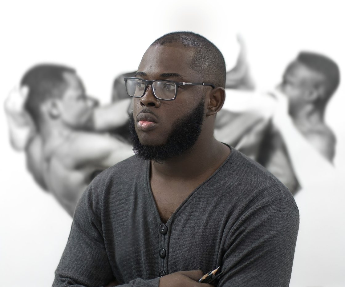 Artist Arinze Stanley wearing glasses in an archival July 2018 portrait, with drawings visible behind him
